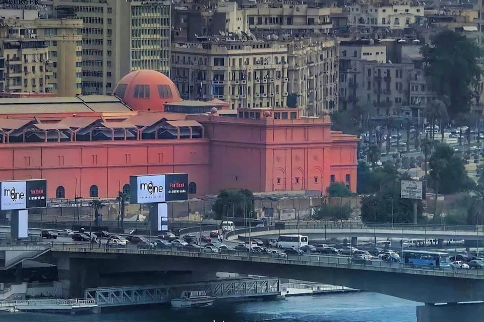 A large orange building and busy bridge in an urban area with heavy traffic and tall beige buildings in the background.