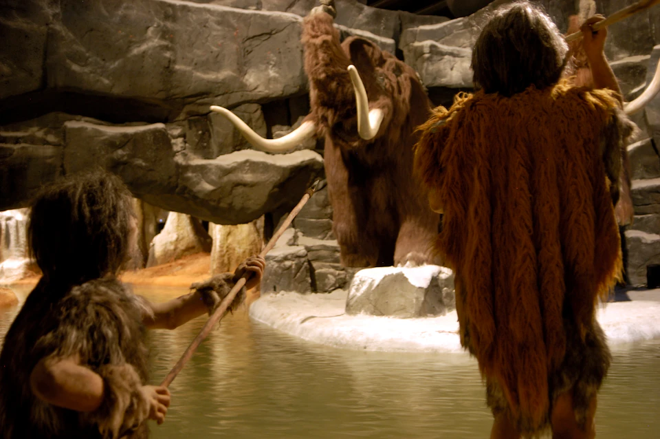 Two figures in fur clothing holding spears face a mammoth near a rocky, water-filled area.