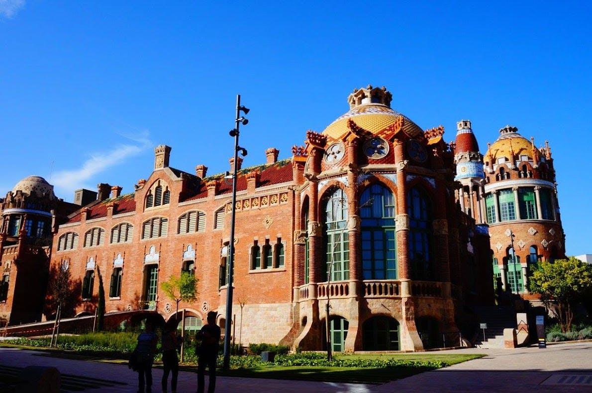 Brick building with ornate dome and arched windows under a clear blue sky; three silhouetted people walking in front.