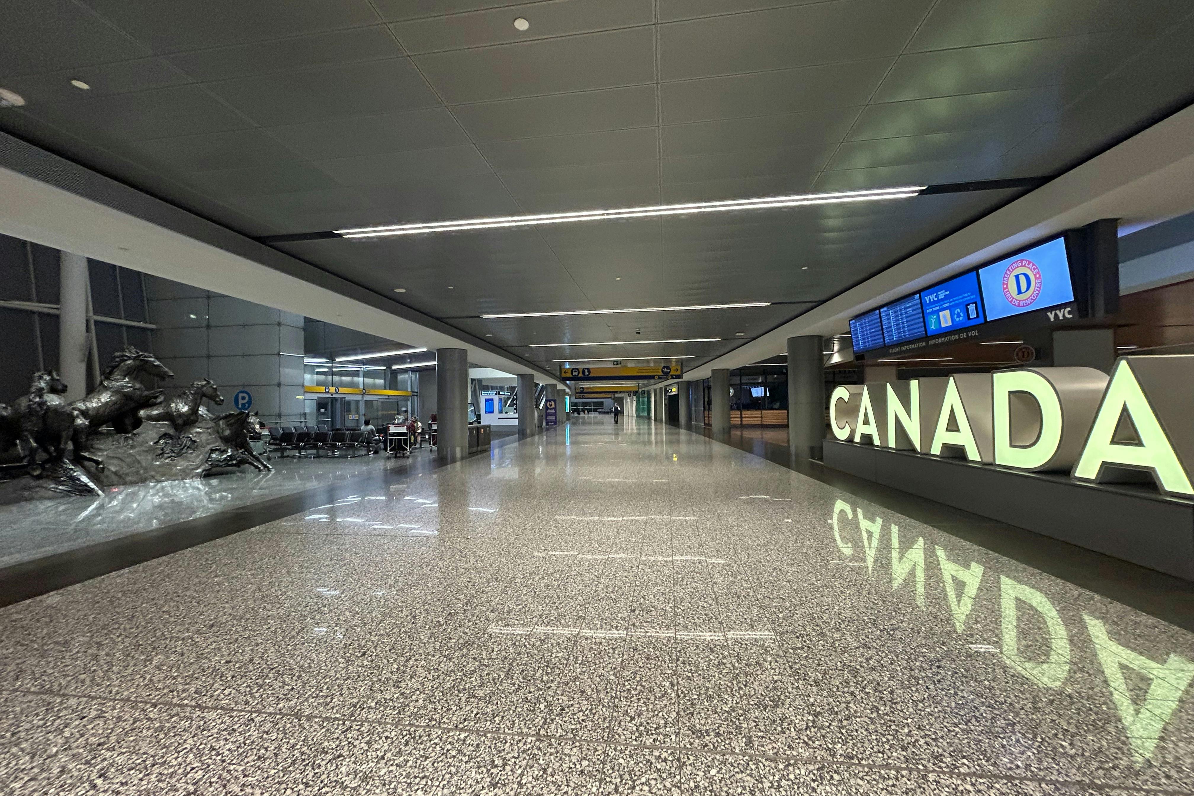 Spacious, modern airport terminal with polished floors, rows of seats, flight information screens, and signs reading "CANADA."