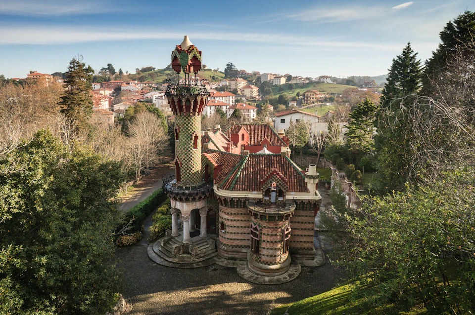 A whimsical, multi-story building with a tiled roof and a decorative tower, set among trees with a village and hills in the background.