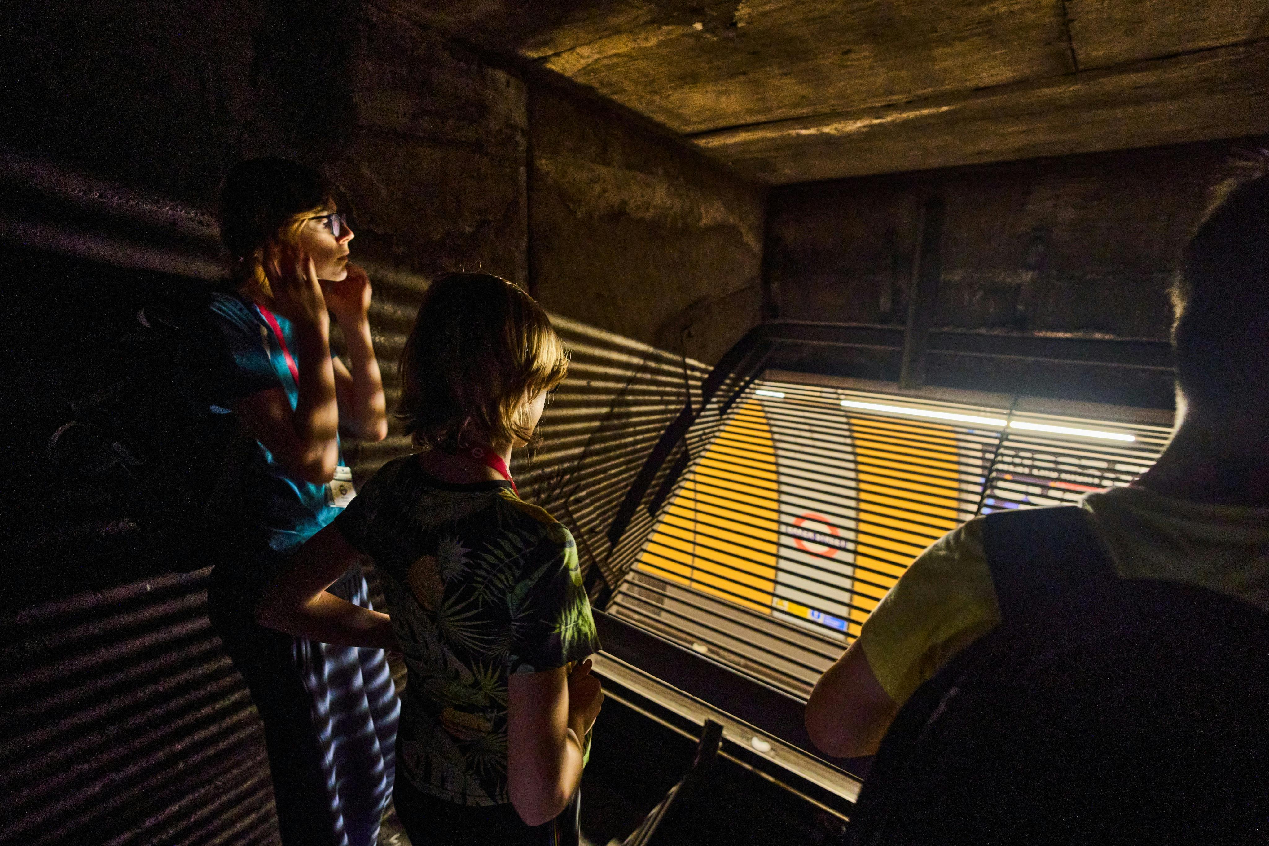 Three people in a dimly lit area looking through a grated barrier into a subway station with a yellow wall.