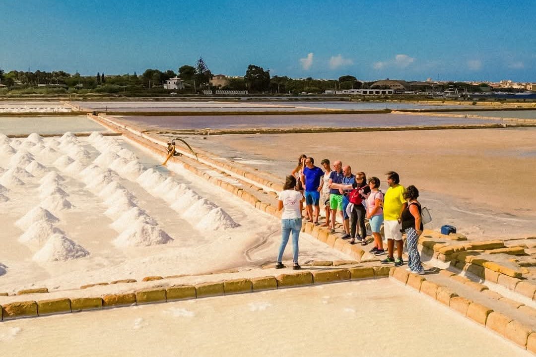 Un gruppo di persone in piedi vicino a cumuli di sale bianco in un'area salina sotto un cielo limpido, con edifici lontani visibili.