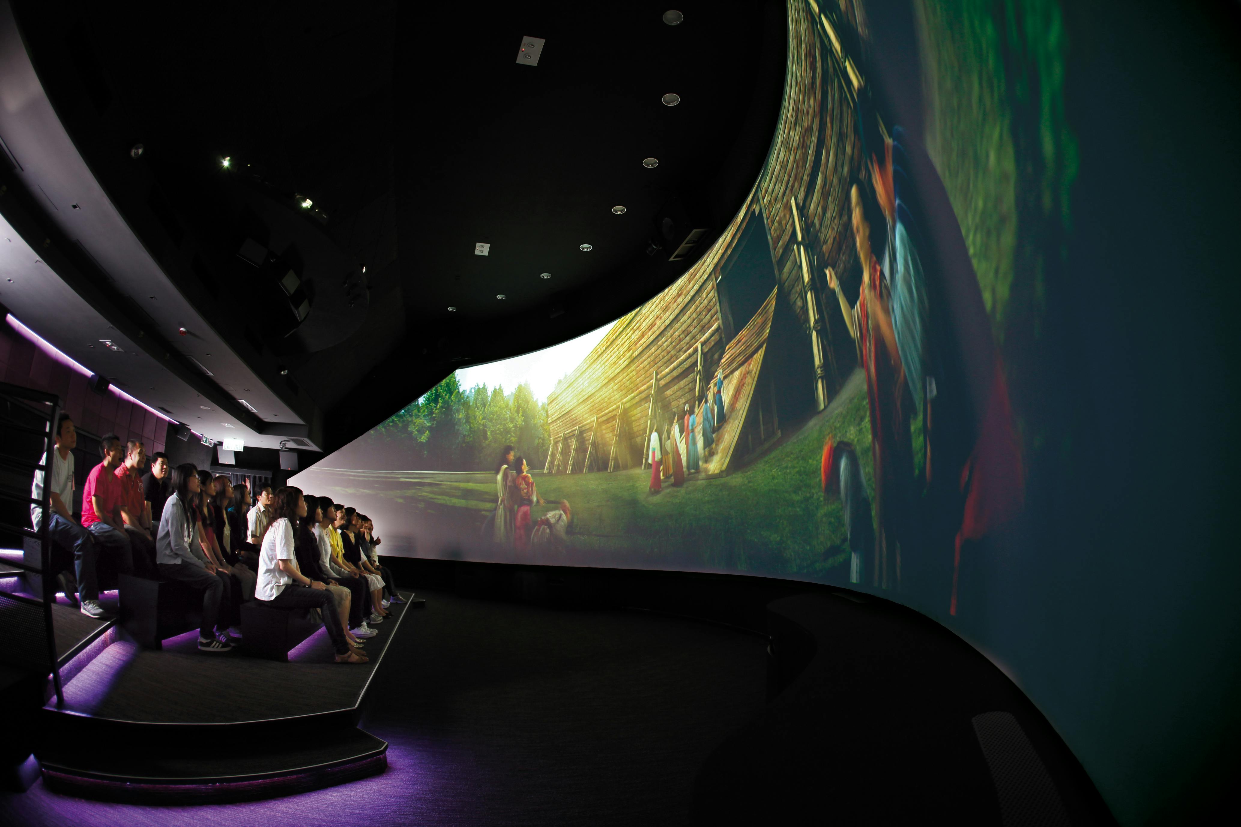 Люди, сидящие в темном кинотеатре, смотрят на большой изогнутый экран, показывающий историческую сцену реконструкции с различными персонажами.