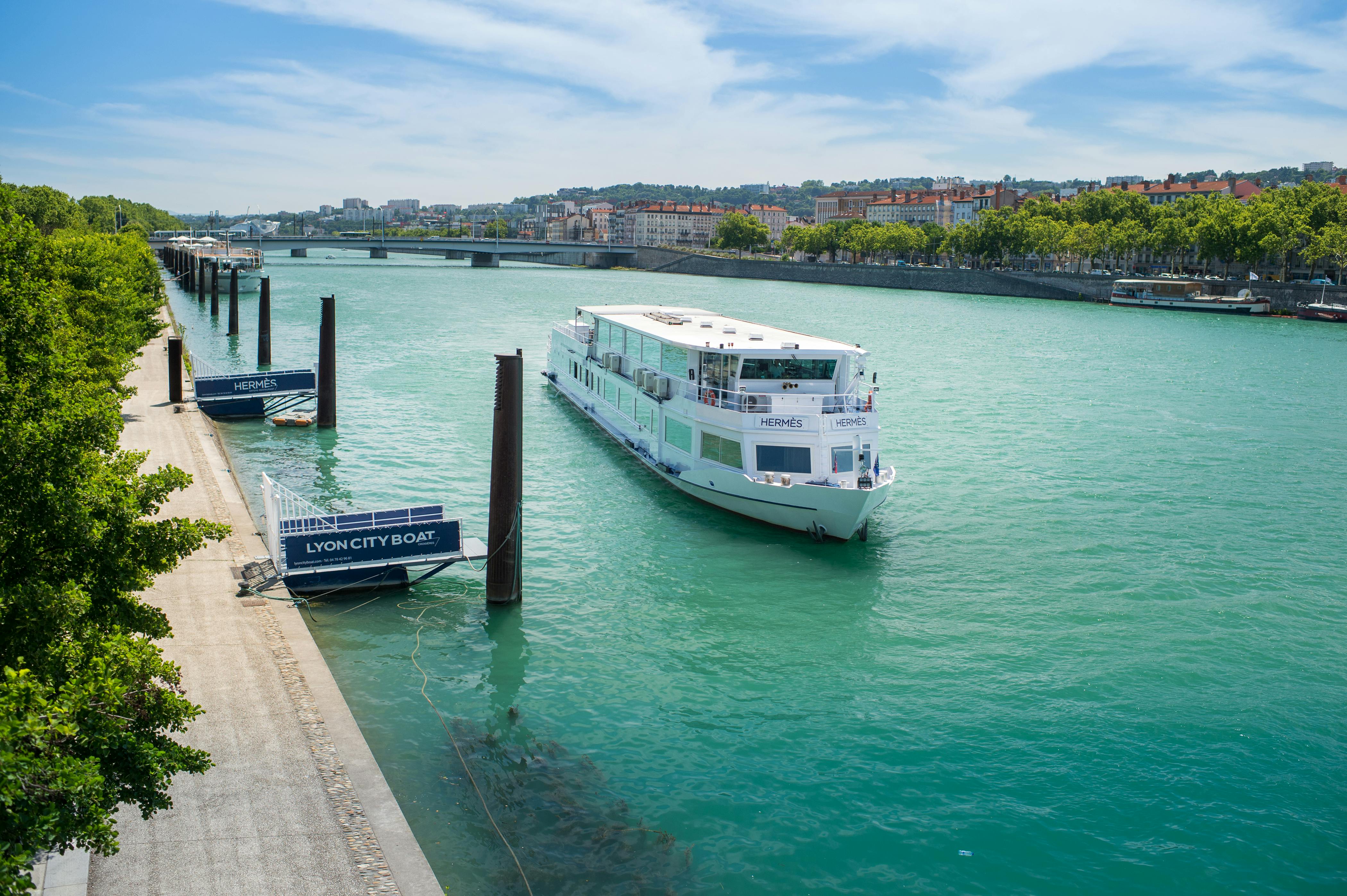 A white cruise boat named "Hermes" is on a wide river near a dock with "Lyon City Boat" signage, surrounded by distant buildings.