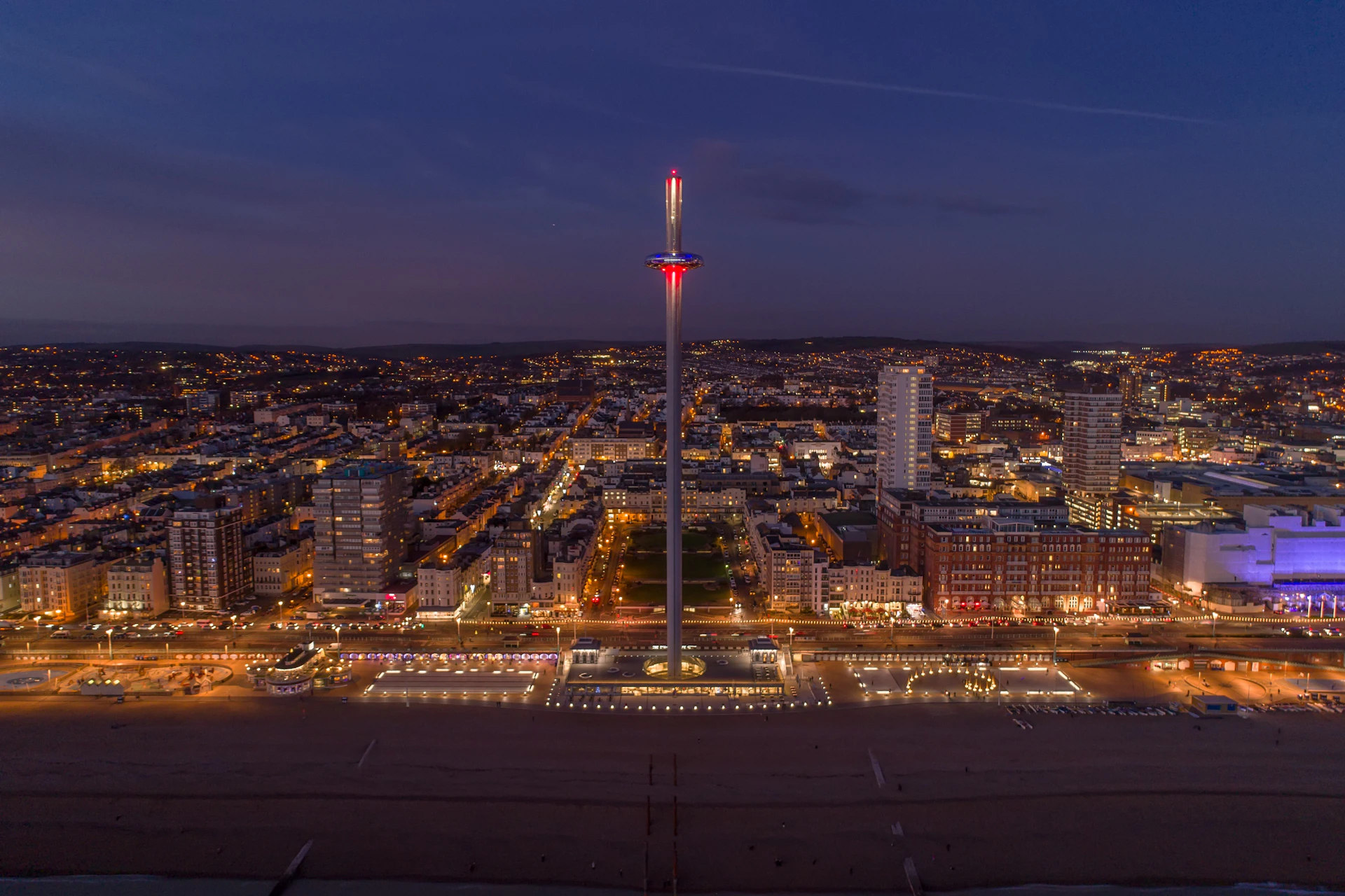 A cityscape at dusk featuring a tall observation tower with lights, surrounded by buildings and illuminated streets.
