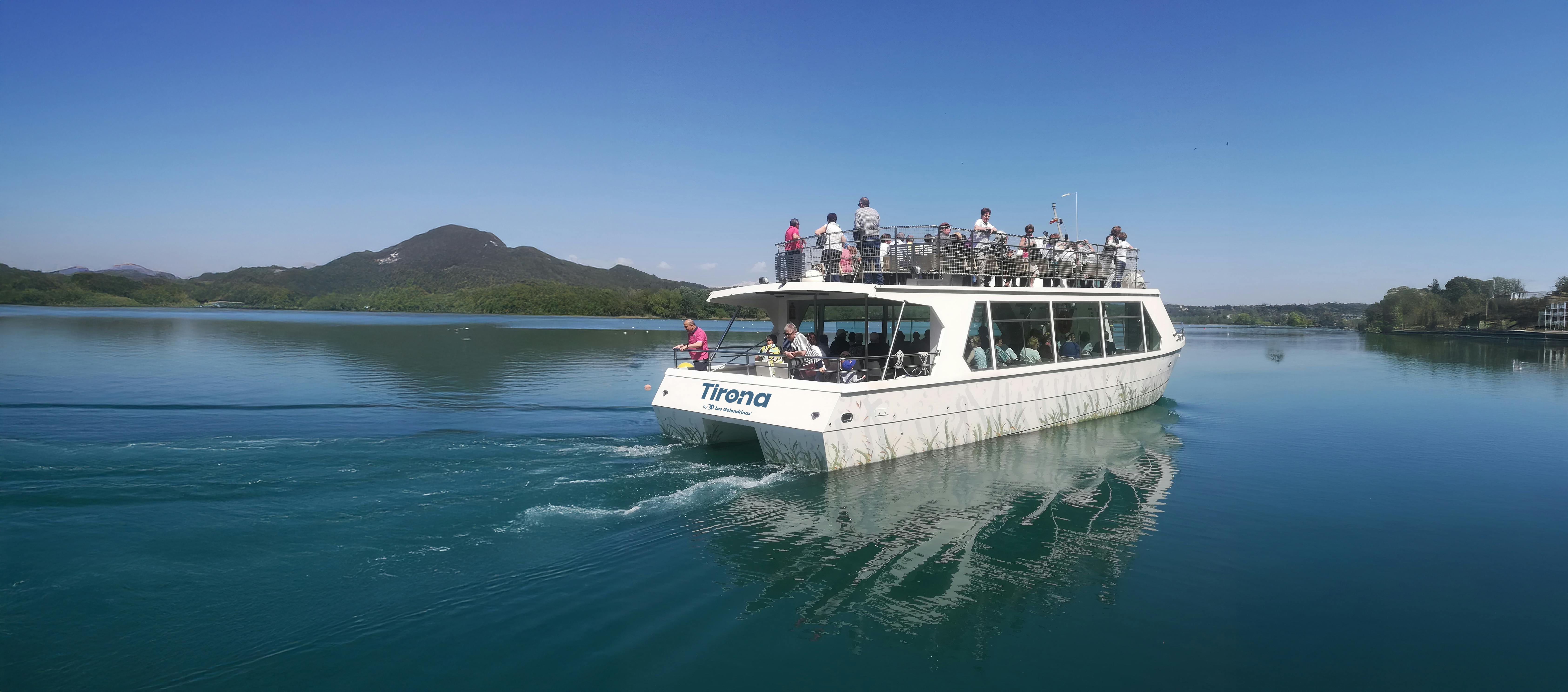 Un vaixell blanc anomenat "Tirona" amb passatgers a les dues cobertes creuant per un llac tranquil amb turons al fons sota un cel blau clar.