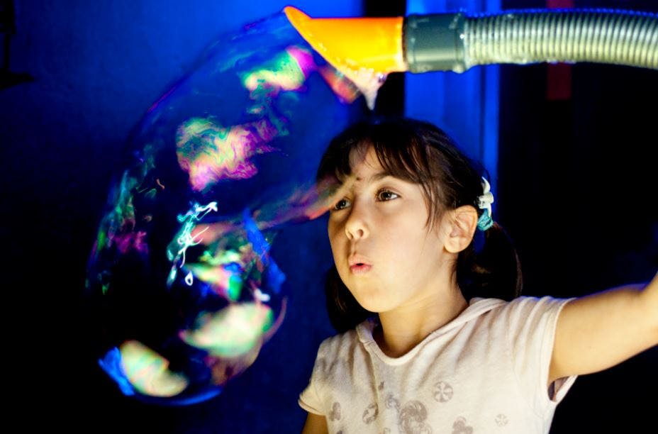 A young girl with pigtails blows a large, colorful bubble using a mechanical device in a dimly lit space.