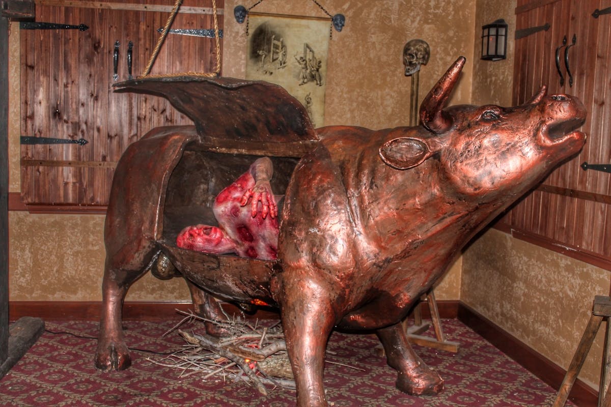 Bronzová socha býka s otevřeným poklopem odhalující uvnitř lidskou postavu, umístěnou v místnosti s dekorem lebky a svitkem na stěně.