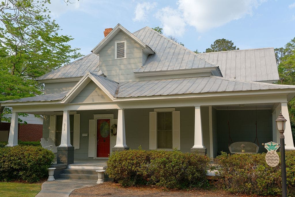 Šedý dům s kovovou střechou, přední verandou, červenými dveřmi a houpačkou. V předzahrádce jsou keře a pouliční lampa.