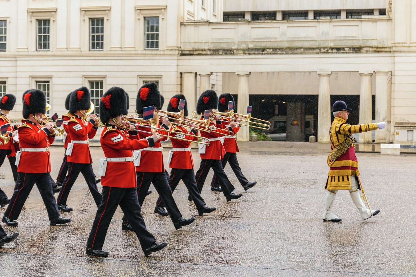 Uniformed marching band in red coats and bearskin hats, playing trombones, passing a ceremonial building with an officer in a yellow uniform.