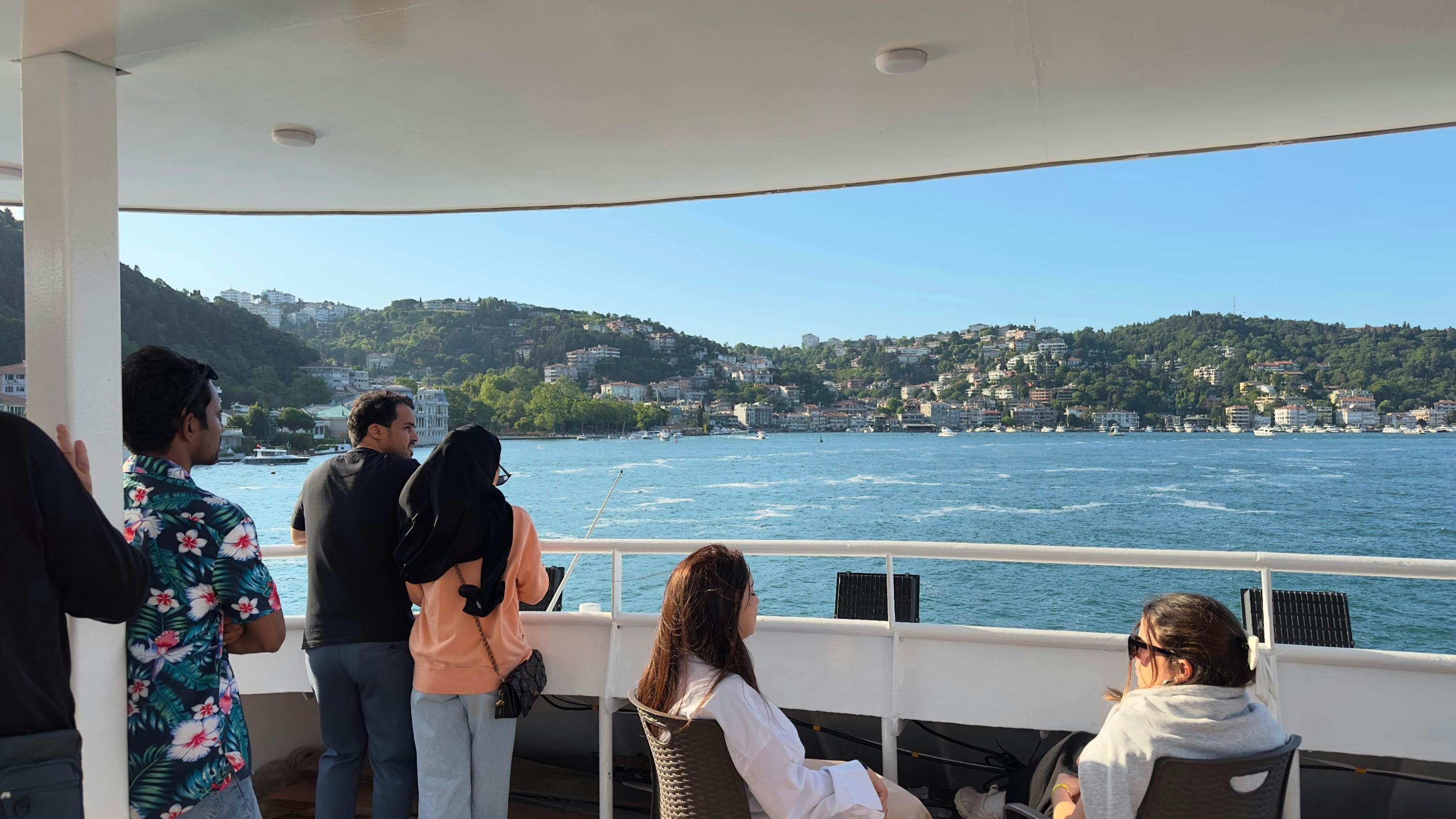 A group of people standing on a boat deck, overlooking a scenic body of water with hills and buildings in the background.
