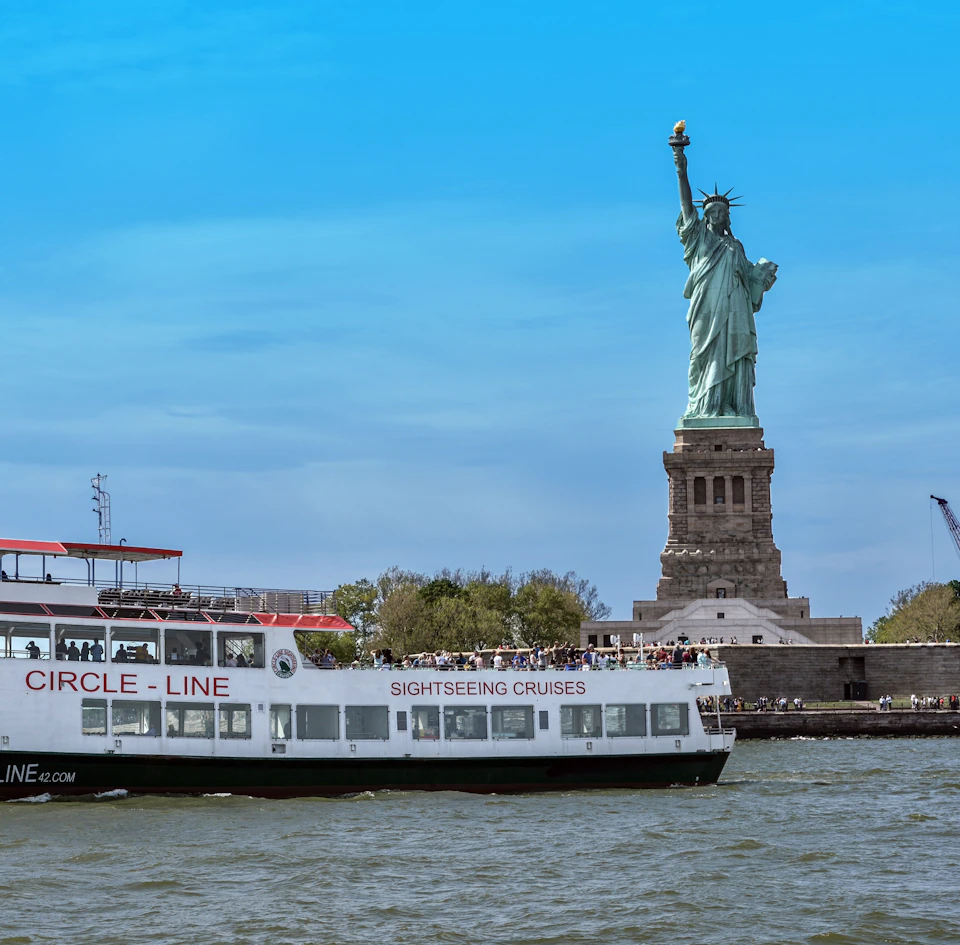 A sightseeing cruise boat passes by the Statue of Liberty on a clear day with a blue sky.