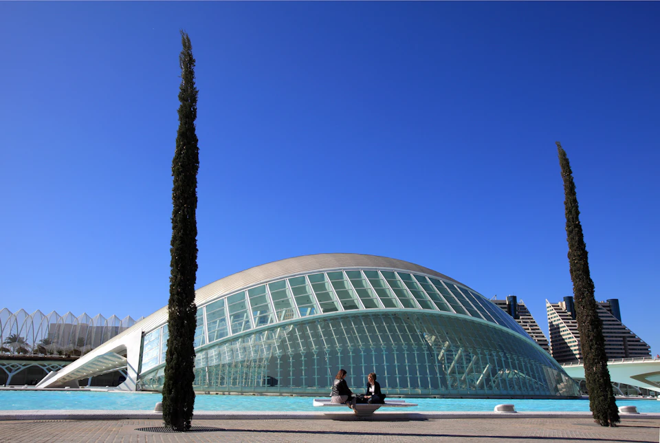 Two people sit on a bench near a large, dome-shaped glass building by a pool, with a tall cypress tree and clear blue sky in the background.