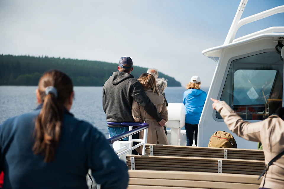 People standing and sitting on a boat, with a lake and tree-covered hills in the background.