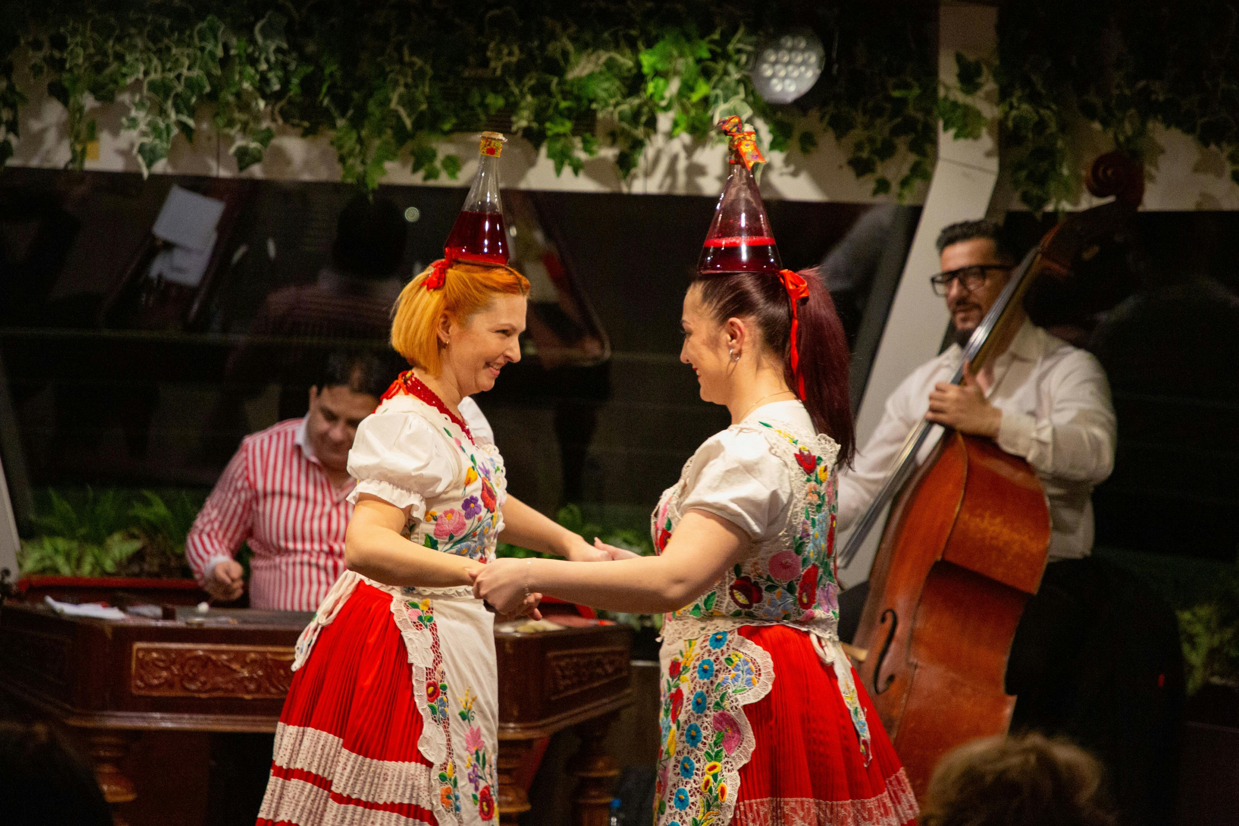Dwie kobiety w tradycyjnym stroju tańczą, noszą czerwono-białe sukienki w kwiatowe wzory i imprezowe kapelusze. W tle gra muzyk.