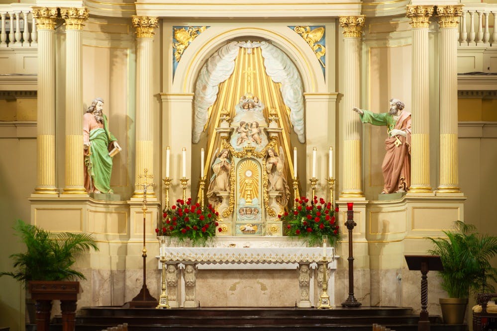 Περίτεχνος βωμός εκκλησίας με αγάλματα, κεριά και κόκκινα λουλούδια. Δύο αγάλματα αγίων πλαισιώνουν την κεντρική μορφή κάτω από ένα χρυσό στέγαστρο.