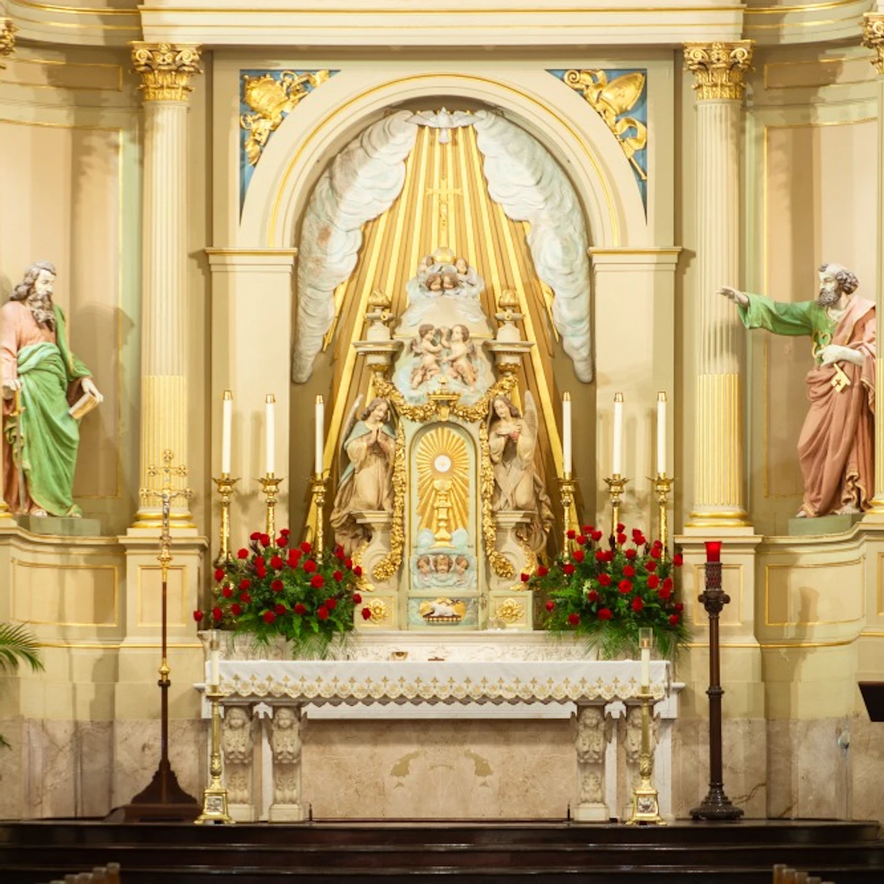 Ornate church altar with statues, candles, and red flowers. Two statues of saints flank the central figure under a golden canopy.