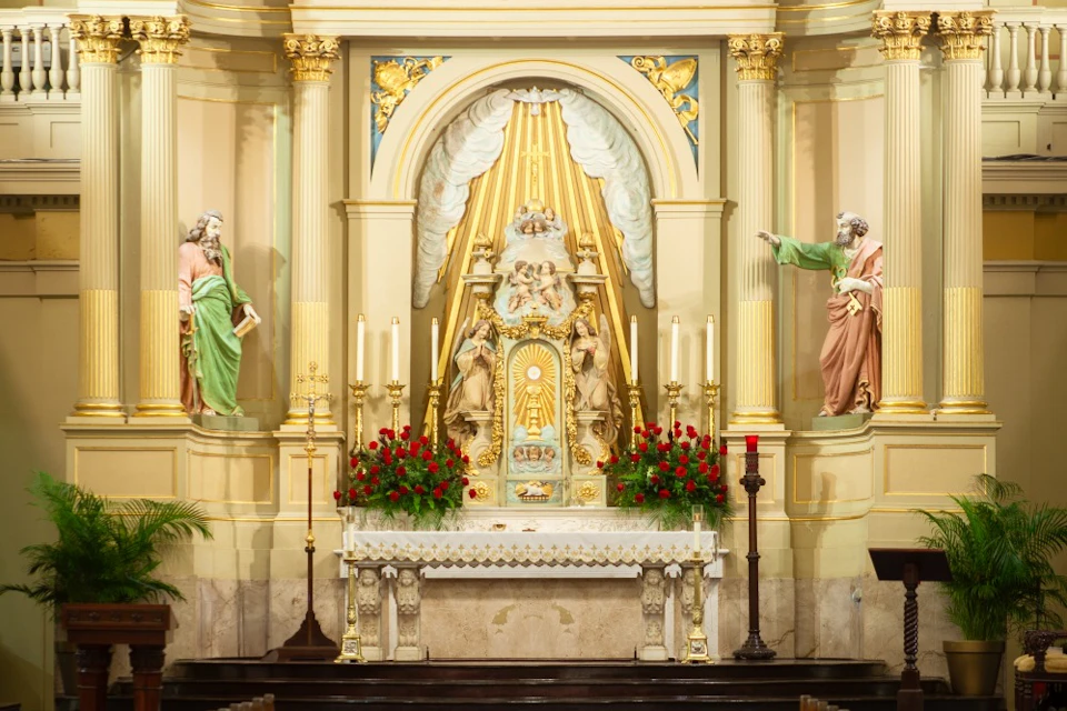 Ornate church altar with statues, candles, and red flowers. Two statues of saints flank the central figure under a golden canopy.
