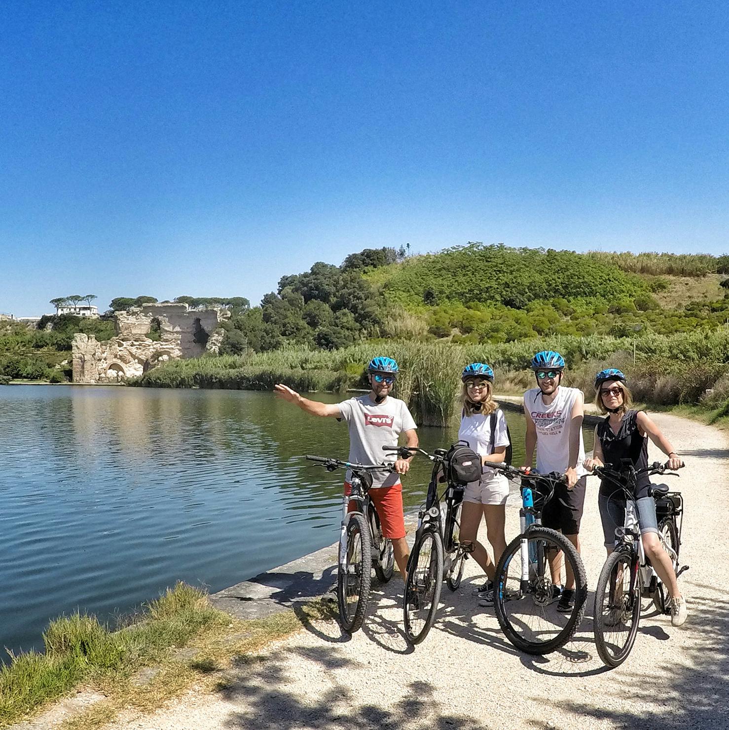 Quattro persone con biciclette e caschi si trovano accanto a un lago con rovine e verde sullo sfondo in una giornata di sole.