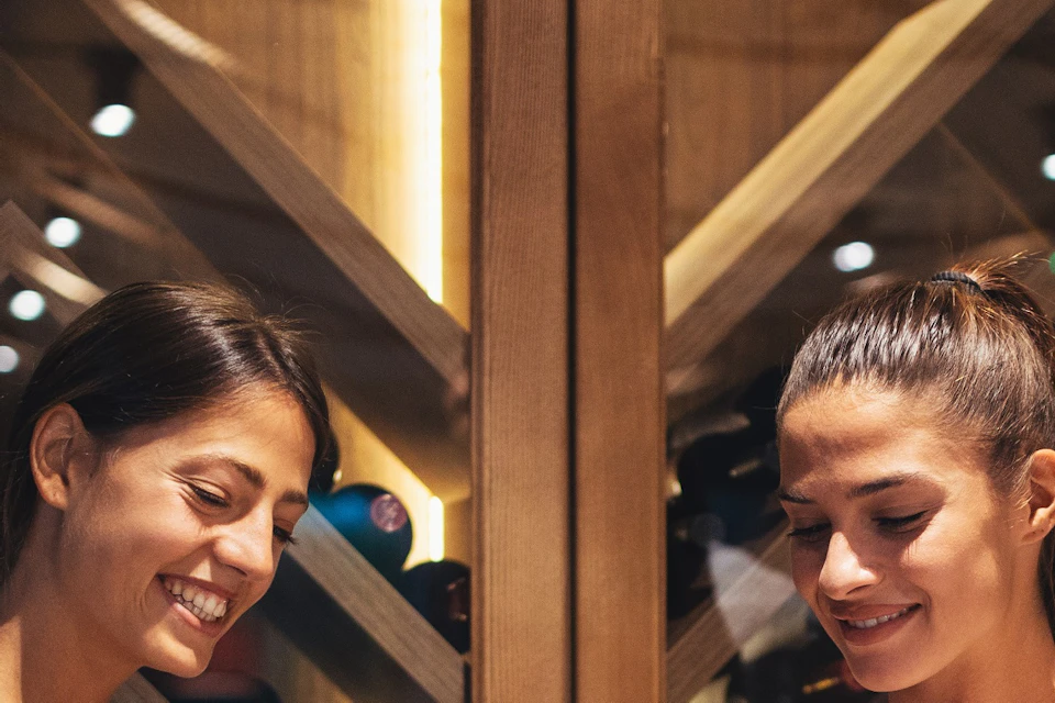 Three people smiling whilst a fourth person pours wine into their glasses in a wine cellar with wooden racks and bottles.