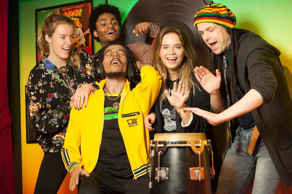 Five people enjoy music together indoors; one plays a drum, another wears headphones. They appear lively and joyful.