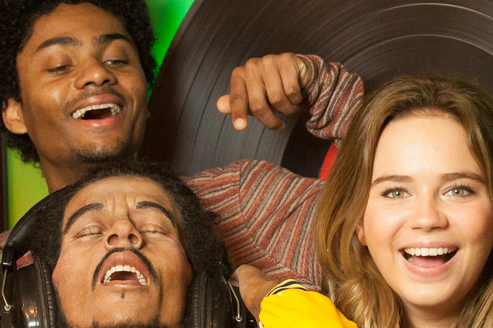 Five people enjoy music together indoors; one plays a drum, another wears headphones. They appear lively and joyful.