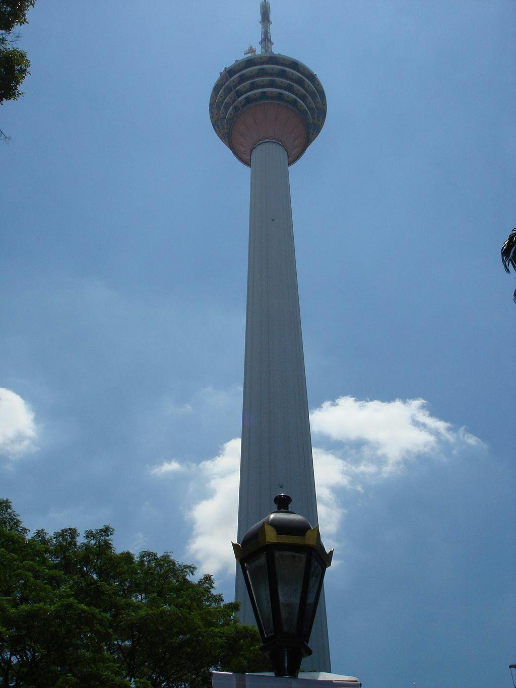 A tall communications tower with a domed top under a cloudy sky, framed by trees and a streetlamp in the foreground.