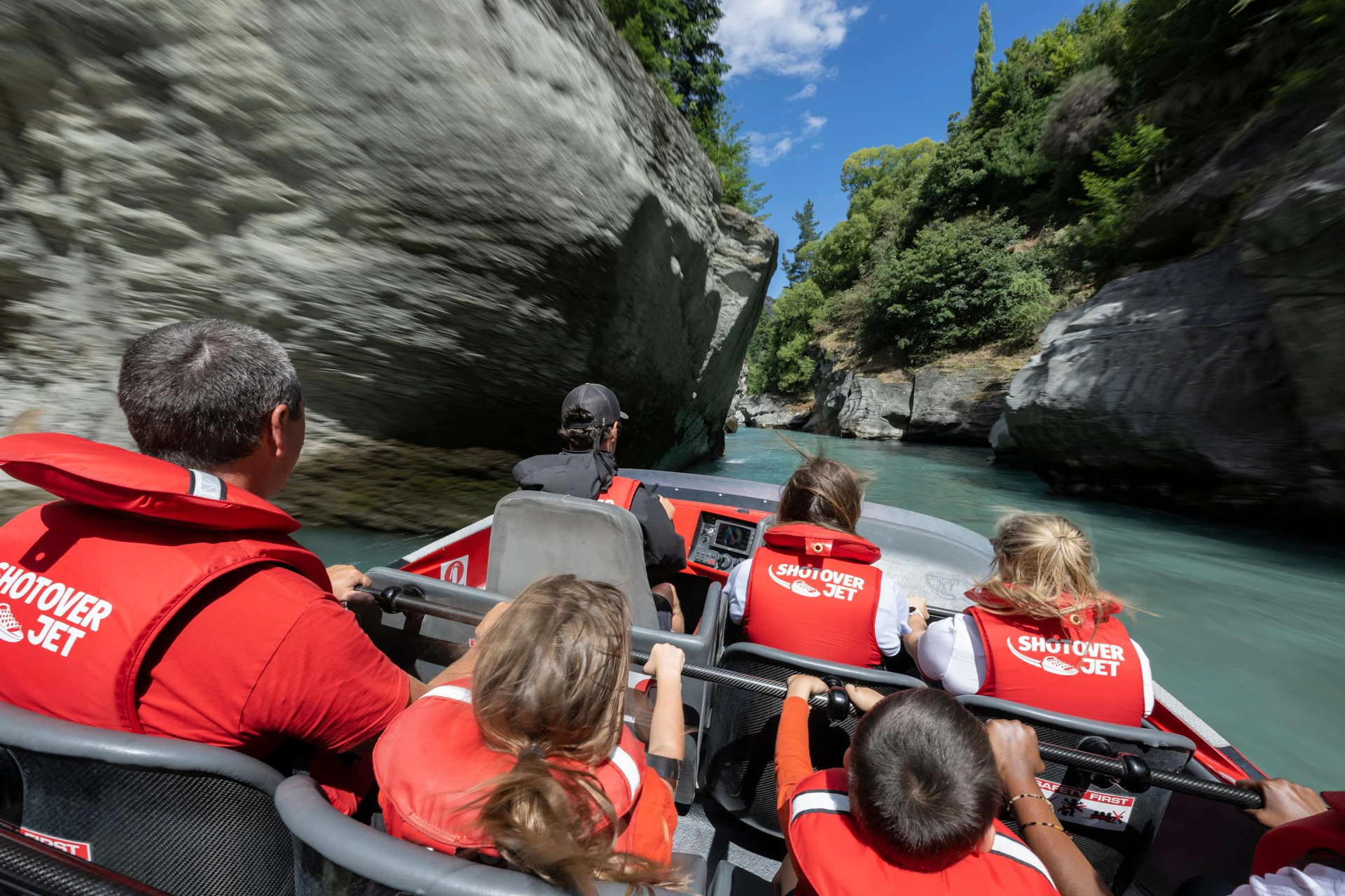 People wearing red life jackets riding in a jet boat through a narrow canyon with rocky walls and trees under a blue sky.