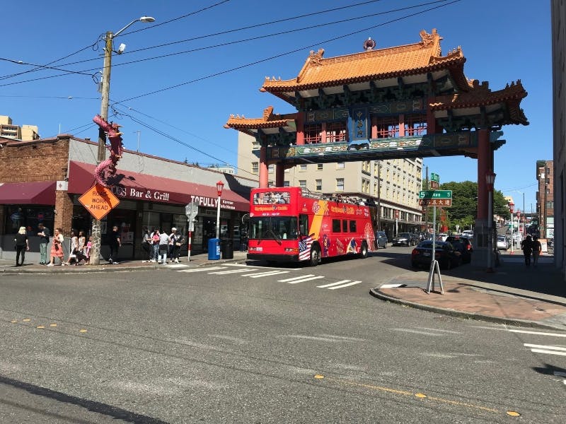 A red double-decker tour bus passes under an ornate, traditional Chinese-style gate in an urban area with shops and pedestrians.