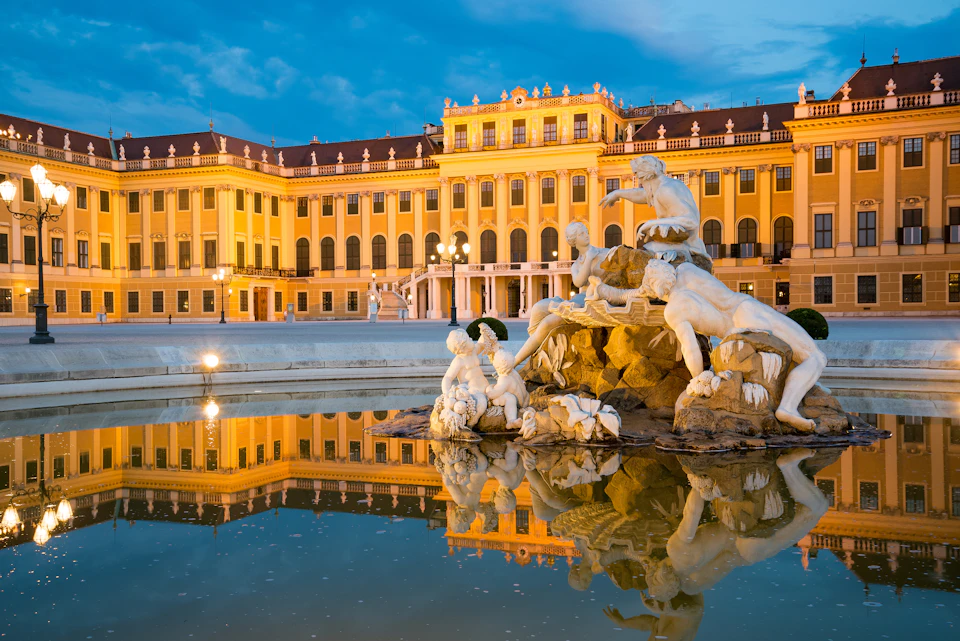 Ornate yellow building with red roof, illuminated at dusk, reflected in a pool beside a detailed white marble statue fountain.