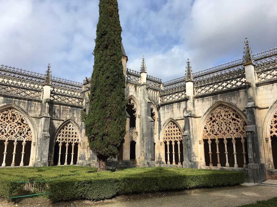 Historic building with intricate Gothic arches and spires, featuring a tall, lush tree and manicured hedges in the courtyard.