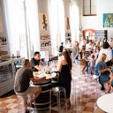 People dining and socializing in a bright restaurant with tall windows, wine bottles on shelves, and paintings on the walls.