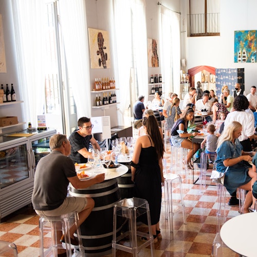 People dining and socializing in a bright restaurant with tall windows, wine bottles on shelves, and paintings on the walls.