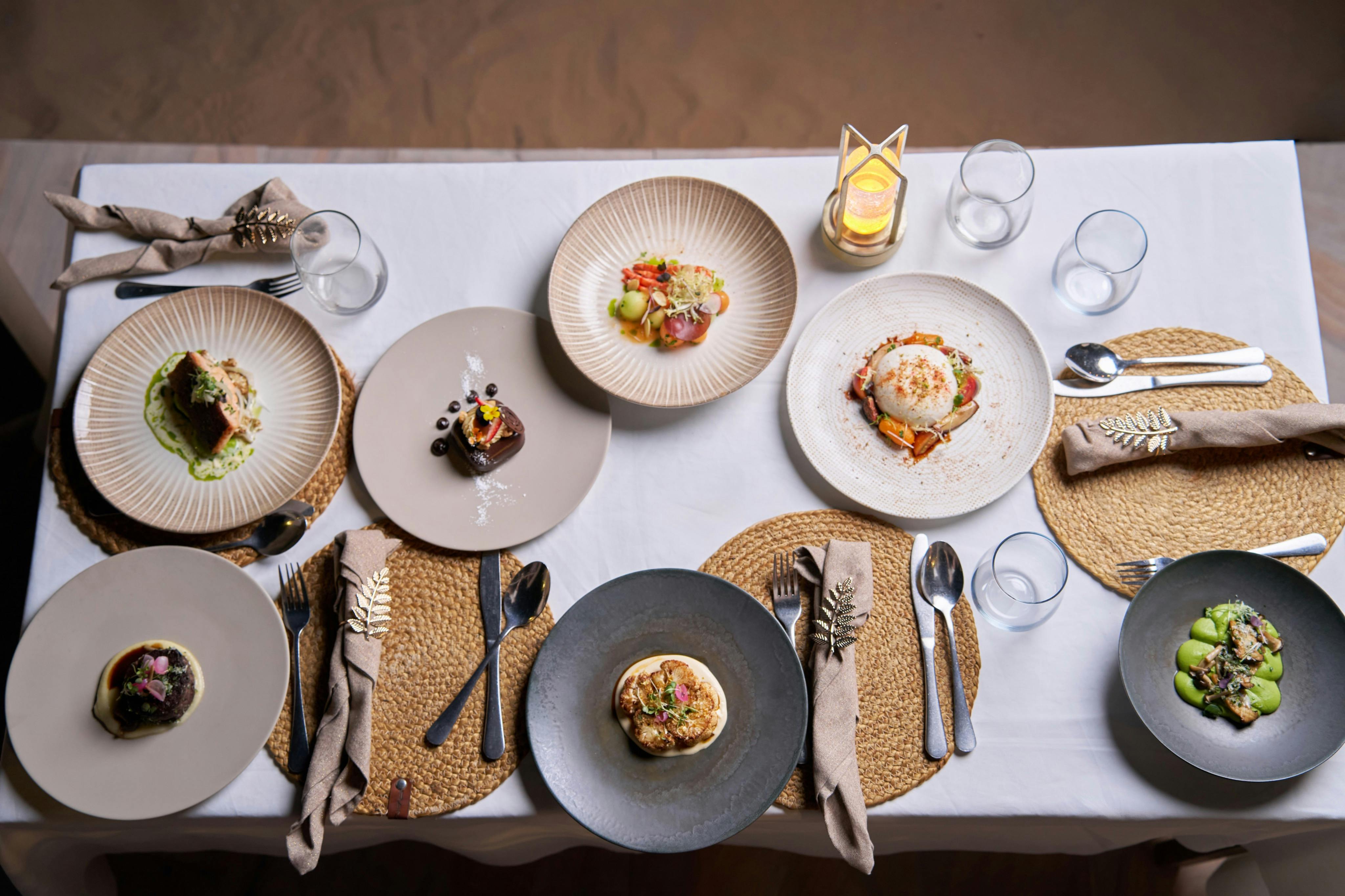 A table set for four with woven placemats, neatly arranged cutlery, napkins, a candle, and plates of gourmet food.