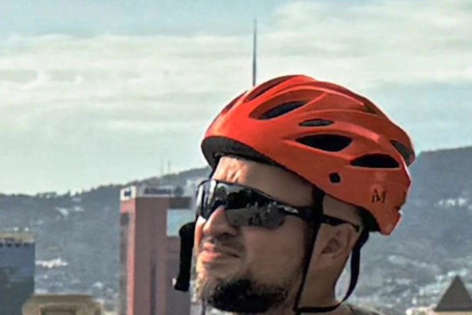 Four people with orange helmets and bikes are outdoors, one pointing at something in the distance, with a cityscape behind.