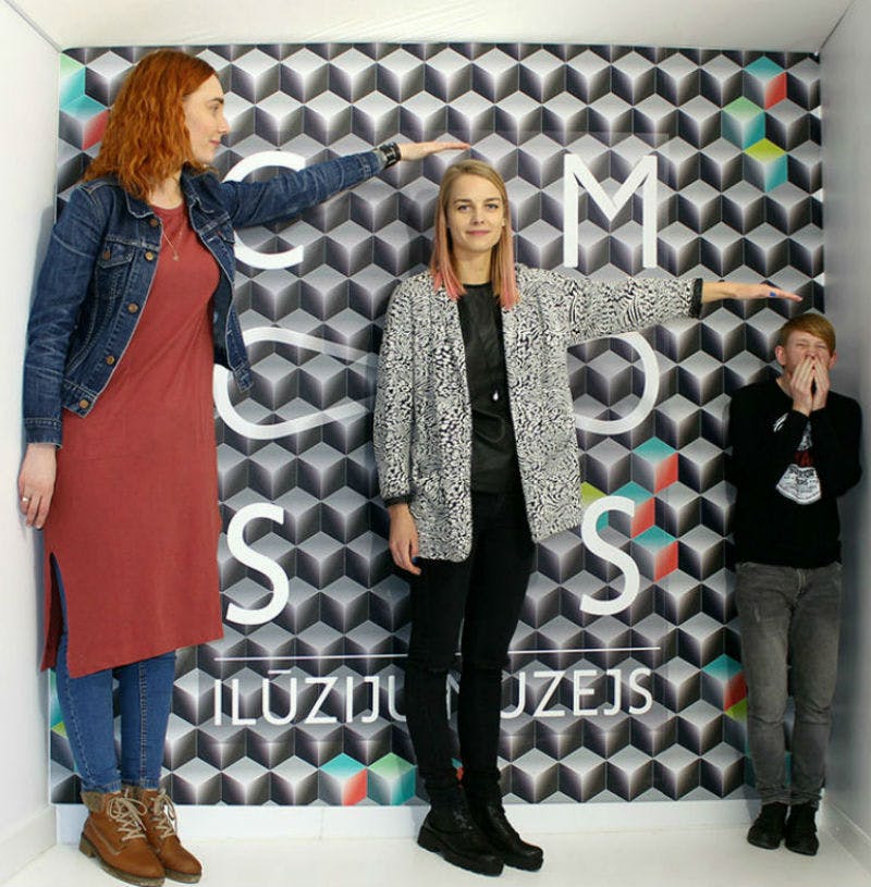 三个人站在一个有视觉错觉的房间里。 背景有几何图案和部分可见的文本。