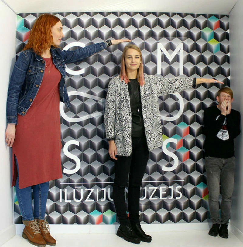 Three people standing in a room with an optical illusion. The background has geometric patterns and partially visible text.