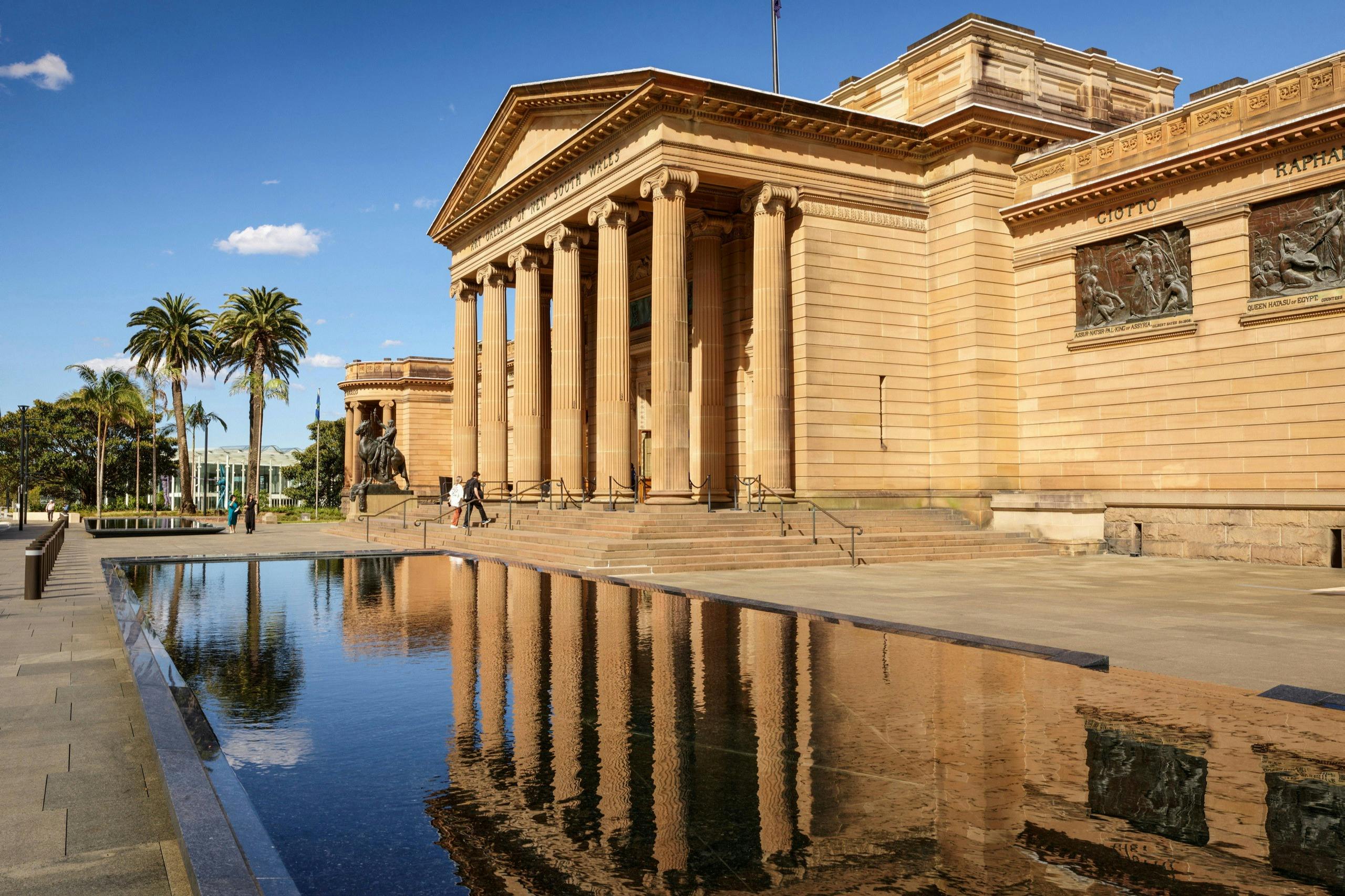 A neoclassical building with tall columns, reflected in a tranquil pool. Two people walk near the entrance. Palm trees and a blue sky are visible.
