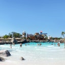 Persone che si divertono in una piscina a onde in un parco acquatico con palme ed edifici sullo sfondo sotto un cielo azzurro e limpido.