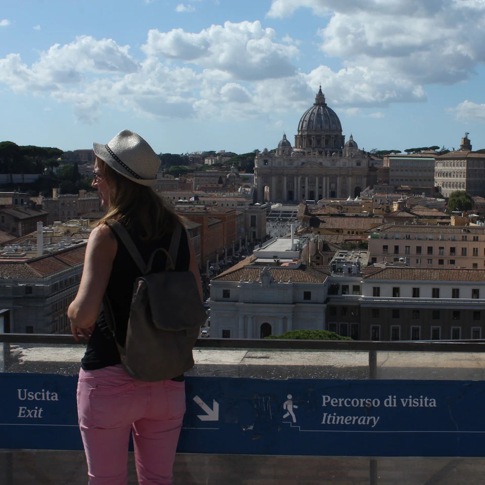 A woman with a backpack overlooks the Vatican City skyline, including St. Peter's Basilica, from a terrace marked with a sign for "Exit" and "Itinerary."