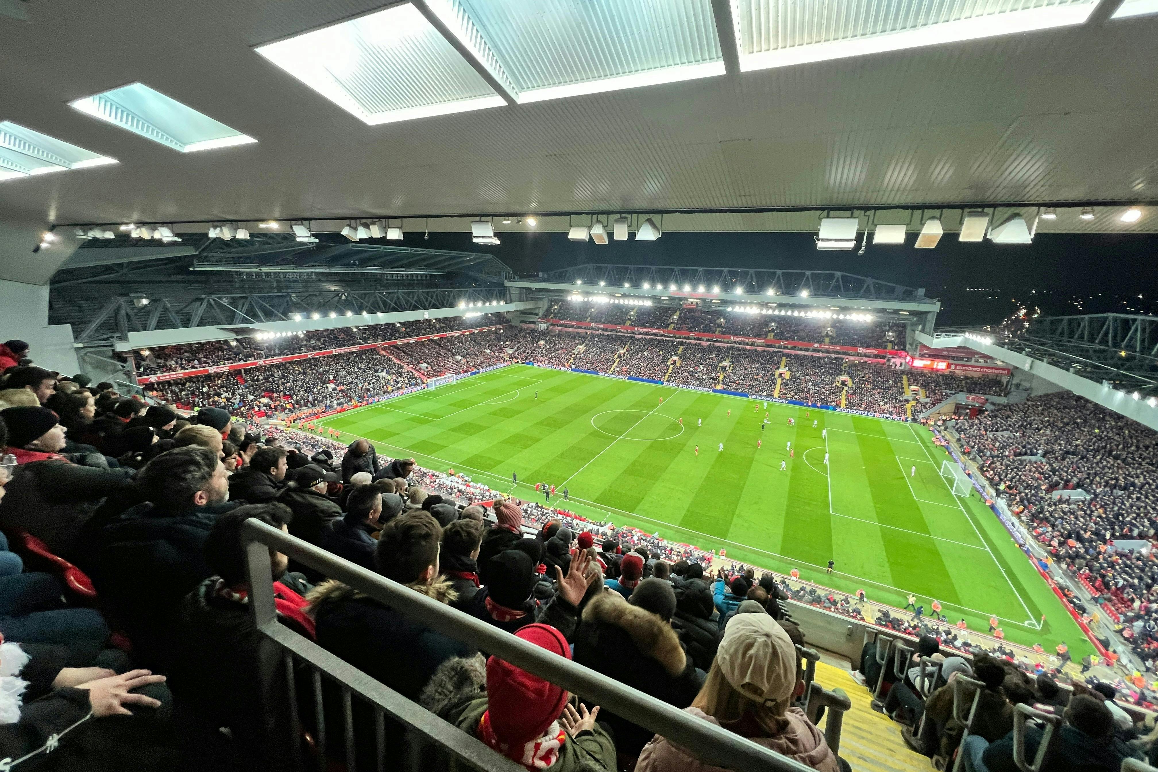 A full stadium watches a nighttime soccer match with fans dressed in winter clothing, viewed from high up in the stands.