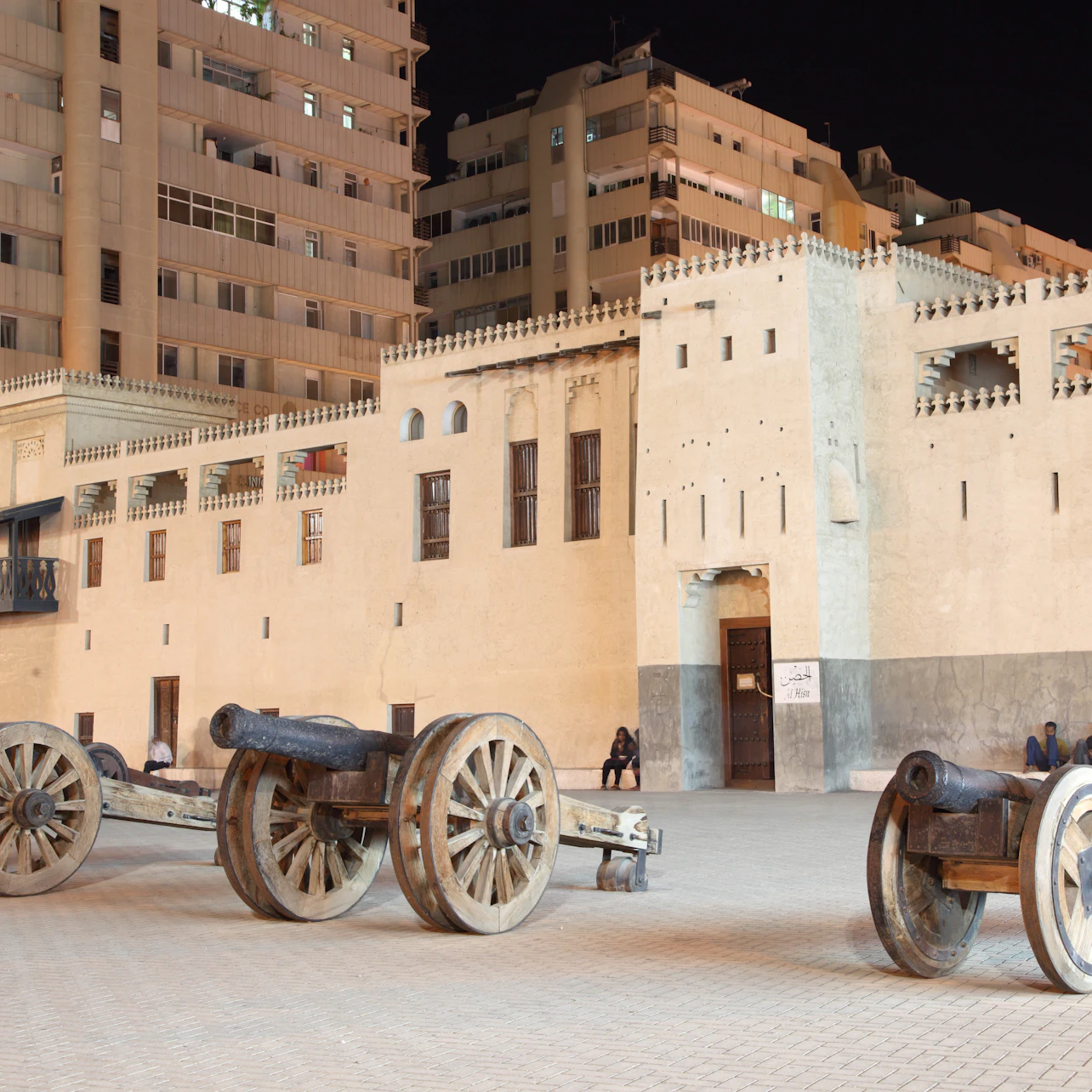 Ancient cannons on wooden wheels displayed in front of a historical building at night, with modern buildings in the background.