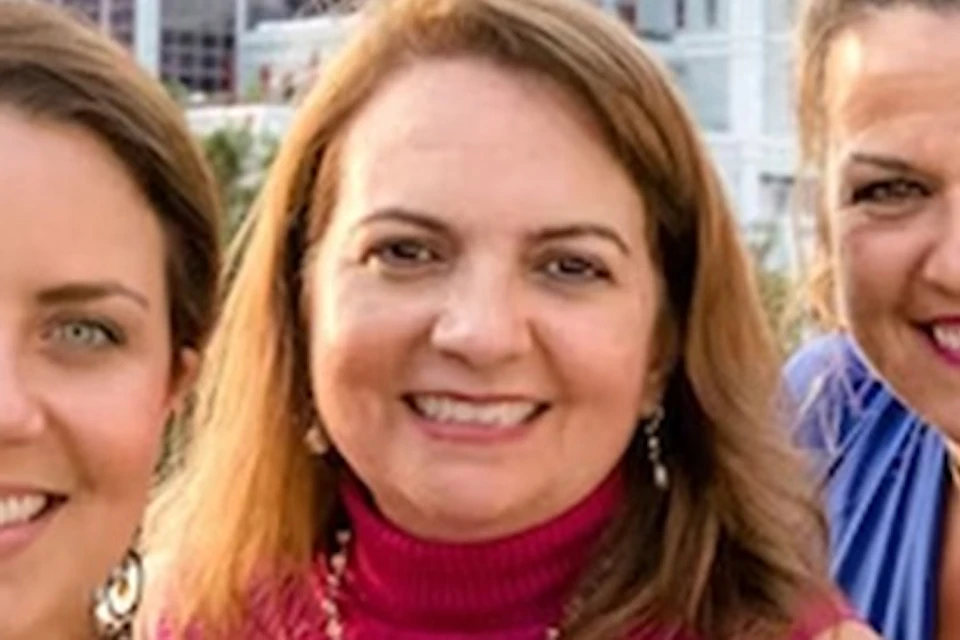 Three women posing outdoors, smiling at the camera, holding drinks, with a background of buildings and trees.