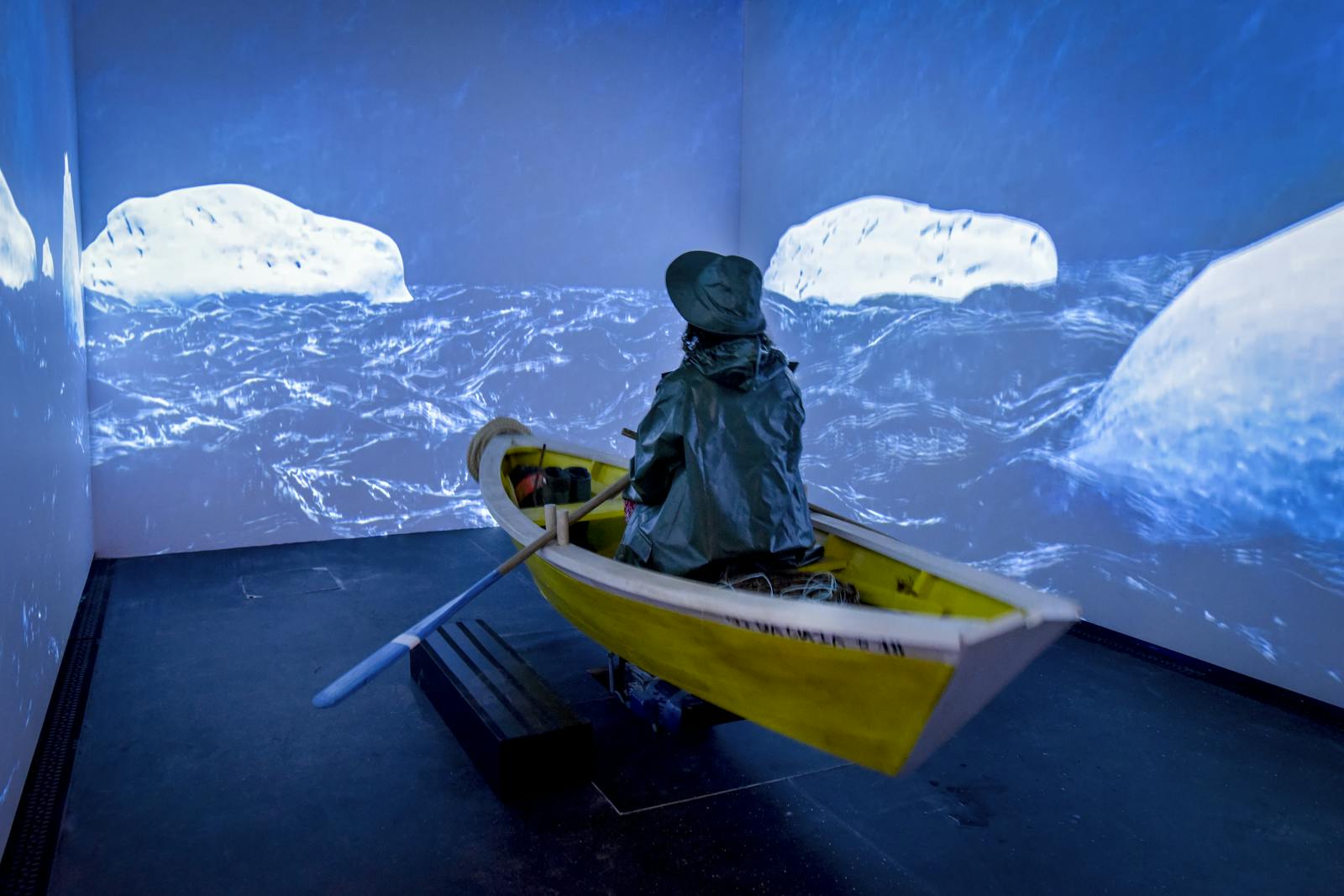 Person in a yellow rowboat, wearing rain gear, appears to row through a virtual seascape projected on surrounding walls.
