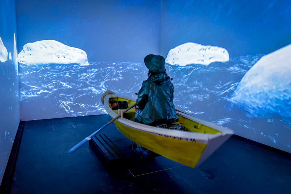 Person in a yellow rowboat, wearing rain gear, appears to row through a virtual seascape projected on surrounding walls.