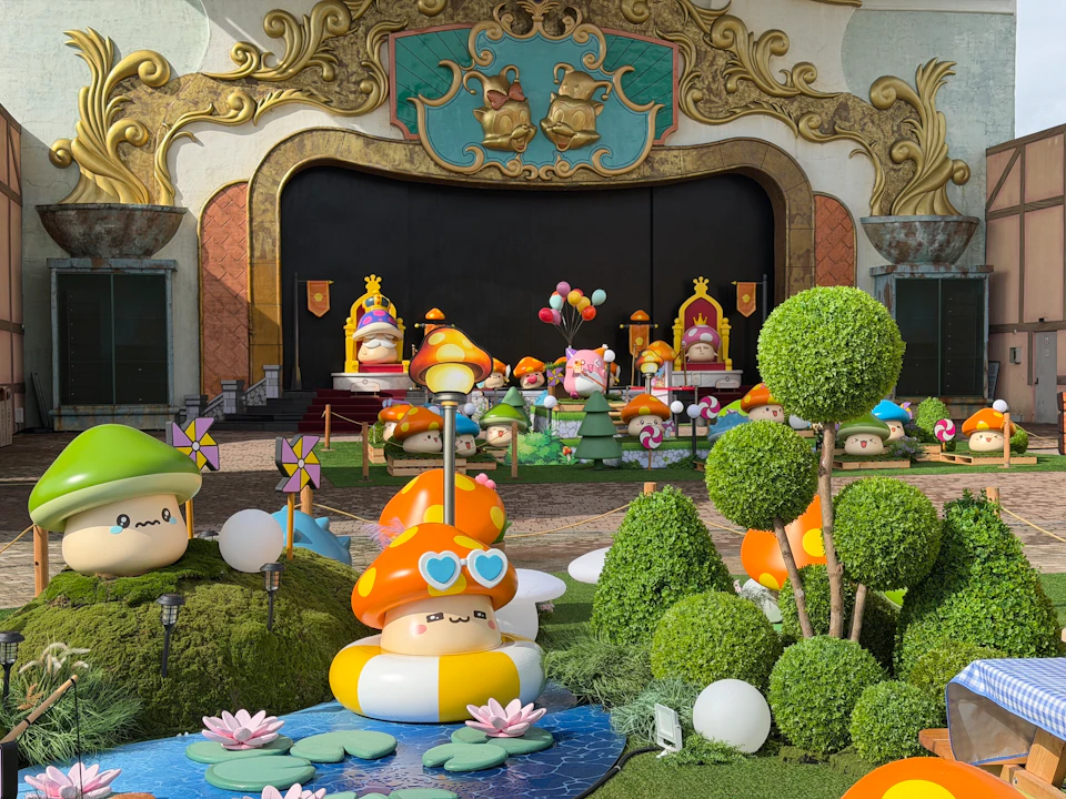 A vibrant outdoor display features colorful mushroom characters, flowers, and decorative elements in front of a grand facade.