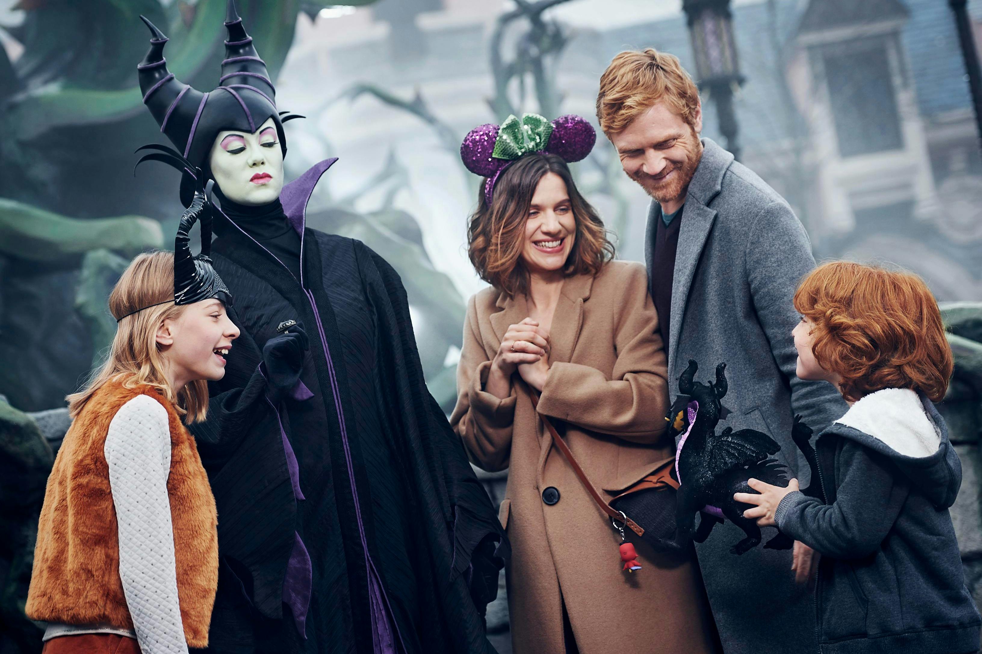 Μια οικογένεια αλληλεπιδρά με ένα άτομο ντυμένο ως Maleficent σε ένα λούνα παρκ, χαμογελώντας και φορώντας διάφορα αξεσουάρ της Disney.