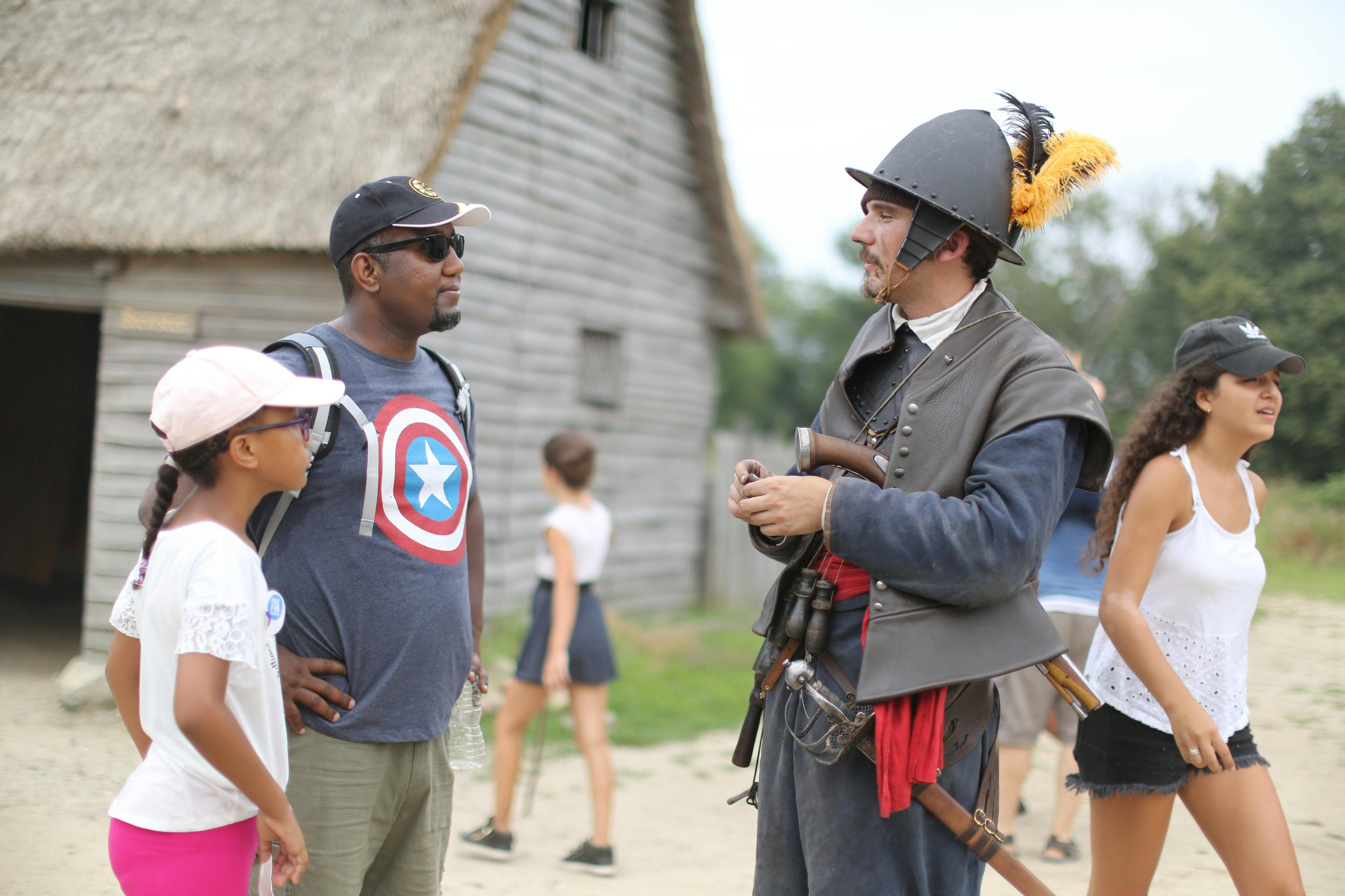 Muž a dítě mluví za slunečného dne s člověkem oblečeným v historickém oděvu poblíž rustikální dřevěné budovy.
