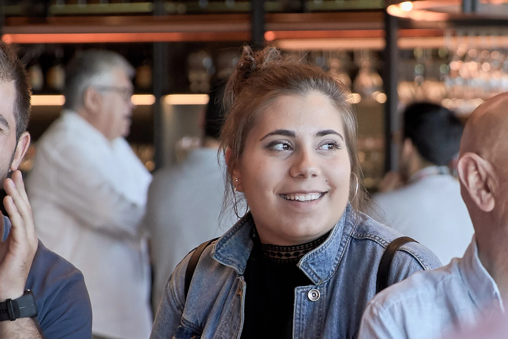 A group of people smile and converse around a wooden table in a modern bar with hanging lights and copper brewing tanks.